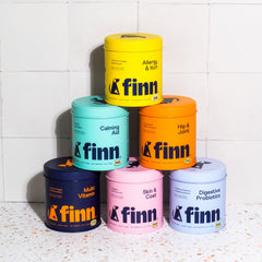 Six colorful Finn pet supplement containers stacked in pyramid formation against white tile wall, featuring different formulas like Calming Aid, Hip & Joint, and Multi Vitamin in bright yellow, teal, orange, navy, pink and purple packaging.
