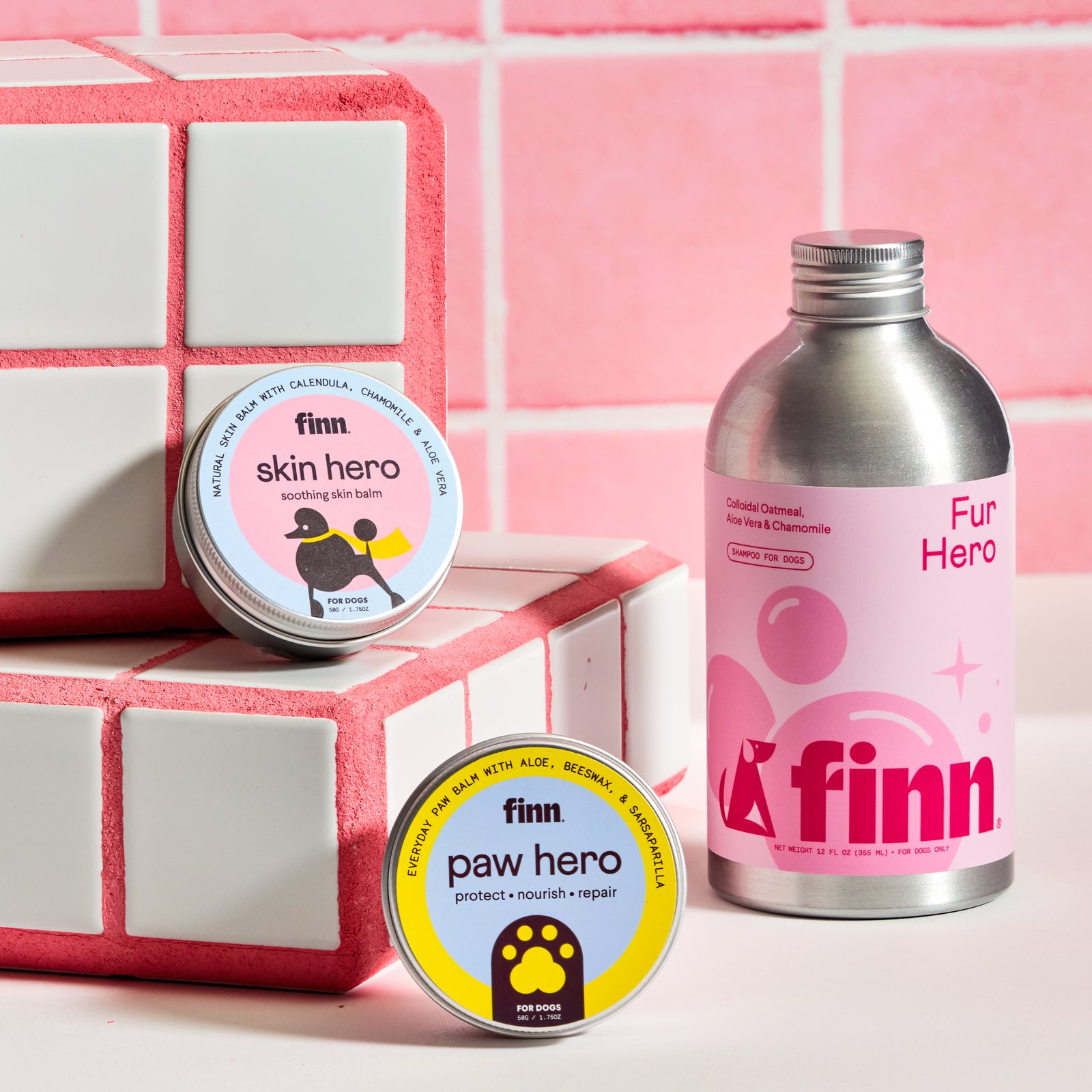 Three Finn dog grooming products on pink tile background: skin hero balm, paw hero balm, and fur hero shampoo bottle
