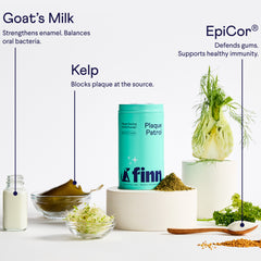 Teal Finn dental powder container surrounded by natural ingredients including goat's milk, kelp, fennel, and herbs on white surface