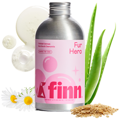 Pink-labeled Finn Fur Hero dog shampoo bottle with silver cap, surrounded by aloe vera leaves, chamomile daisies, cucumber slices, and oats on black background.