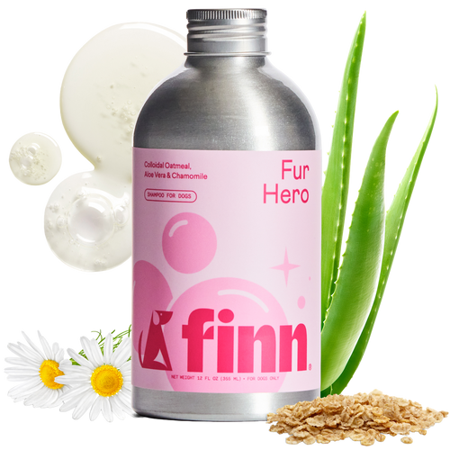 Pink-labeled Finn Fur Hero dog shampoo bottle with silver cap, surrounded by aloe vera leaves, chamomile daisies, cucumber slices, and oats on black background.
