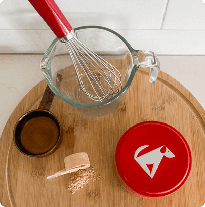 Glass mixing bowl with red-handled whisk on wooden cutting board, alongside small bowls and red container with dog logo for baking pet treats