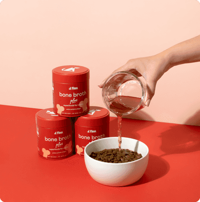 Hand pouring bone broth powder from glass jar into white bowl, with three red cylindrical containers stacked nearby on coral background.