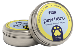 Two round metal tins of Finn paw hero dog paw balm with yellow and blue labels featuring paw print logos, one tin open showing the product inside.