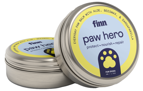 Two round metal tins of Finn paw hero dog paw balm with yellow and blue labels featuring paw print logos, one tin open showing the product inside.