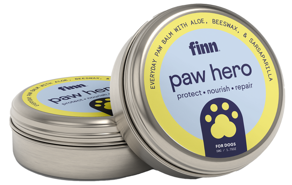 Two round metal tins of Finn paw hero dog paw balm with yellow and blue labels featuring paw print logos, one tin open showing the product inside.