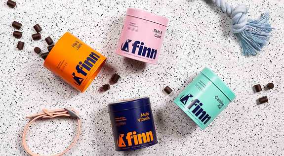 Four colorful Finn supplement containers in orange, pink, mint green, and dark blue arranged on speckled white surface with scattered dark cubes and rope accessories.