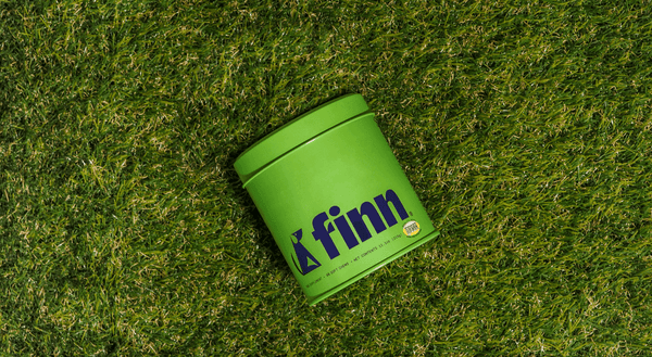 Green cylindrical tin container with "finn" branding in purple text, sitting on artificial grass turf