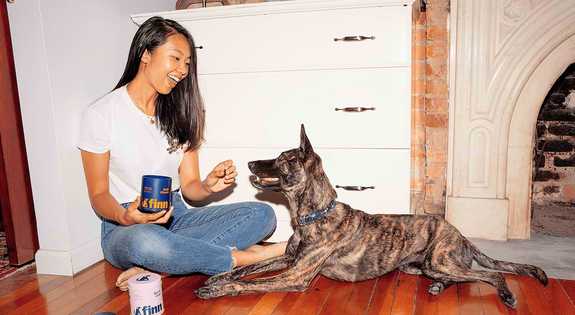 Woman in white shirt sits on wooden floor feeding treats to brindle dog beside white cabinet