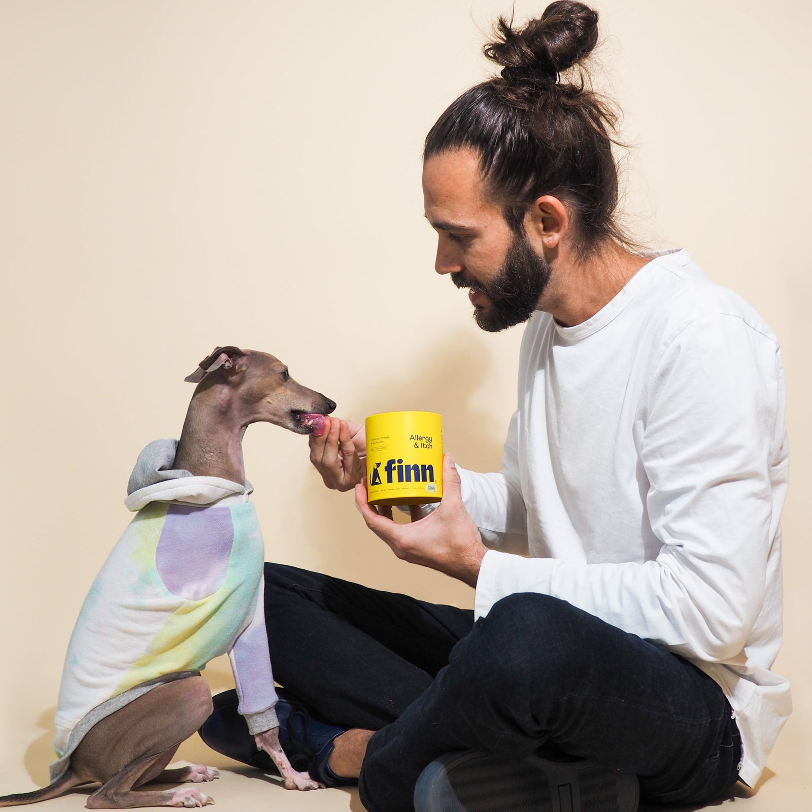 Man in white shirt sits cross-legged feeding treat to dog wearing colorful tie-dye hoodie, holding yellow FINN container