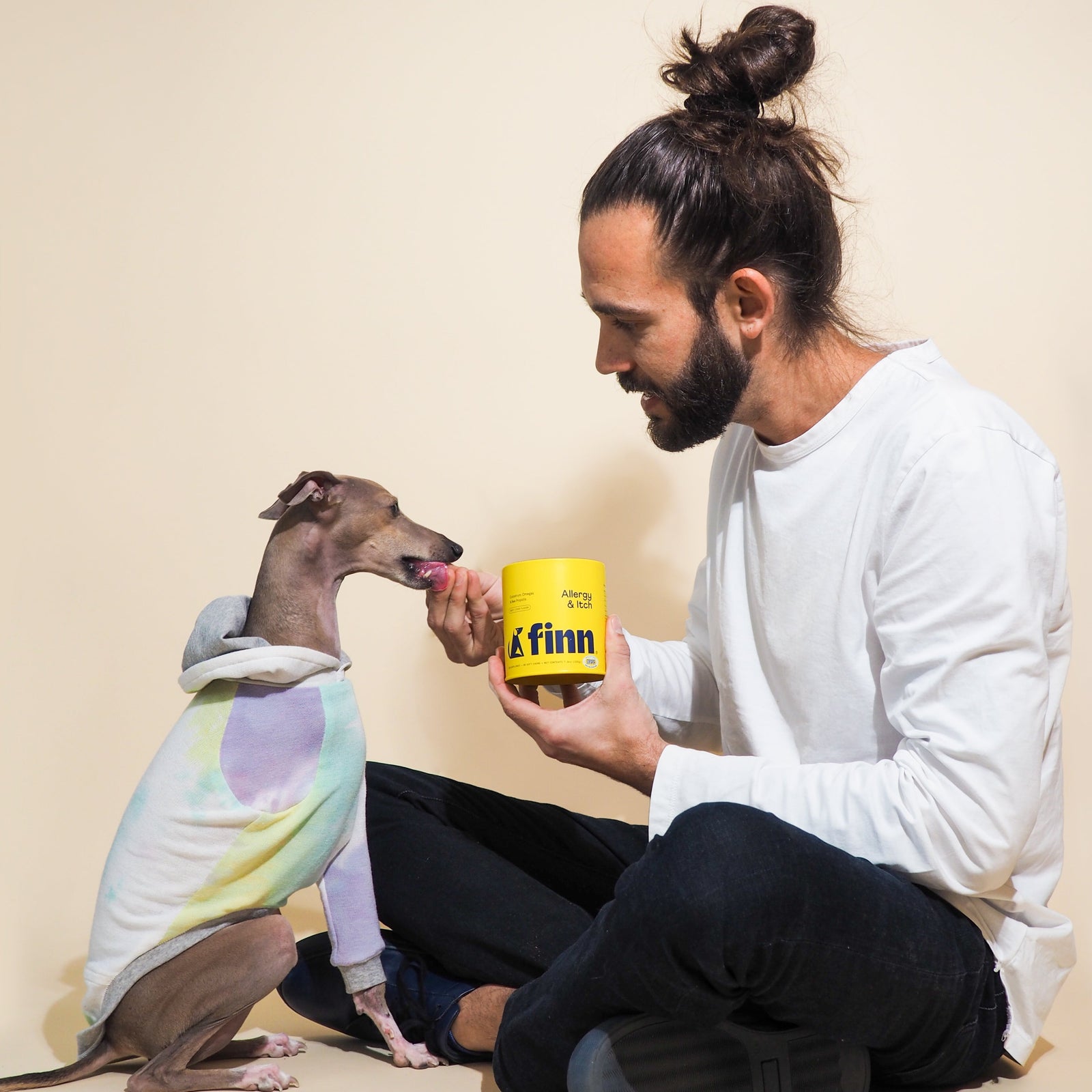 Man in white shirt feeding treat from yellow FINN container to small dog wearing colorful tie-dye hoodie