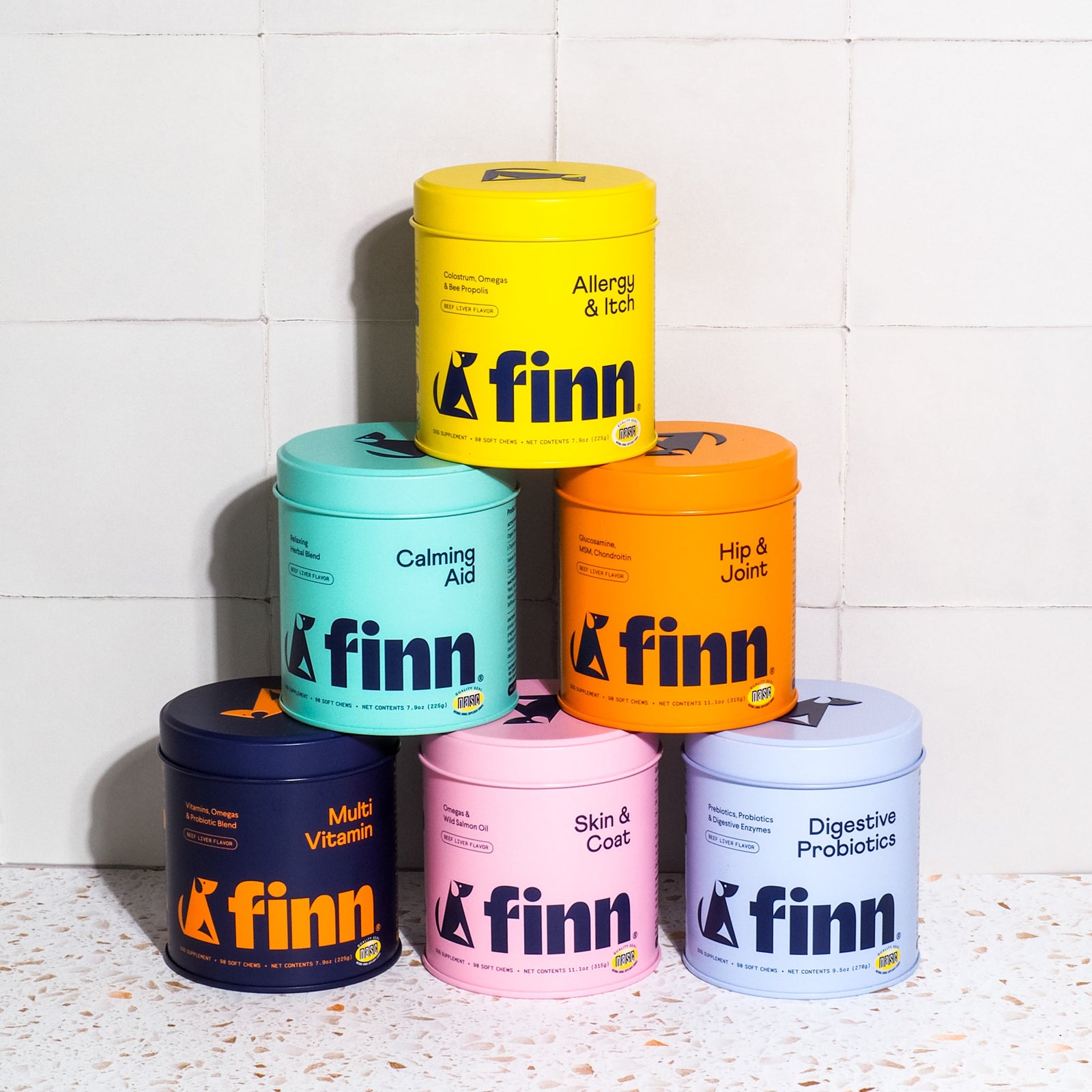 Six colorful Finn pet supplement containers stacked in a pyramid - yellow, teal, orange, navy, pink, and light blue - against white tile wall.