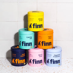 Six colorful Finn pet supplement containers stacked in a pyramid - yellow, teal, orange, navy, pink, and light blue - against white tile wall.