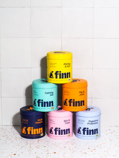 Stack of six colorful Finn pet supplement containers in yellow, teal, orange, navy, pink, and light blue arranged in pyramid formation against white tile wall.