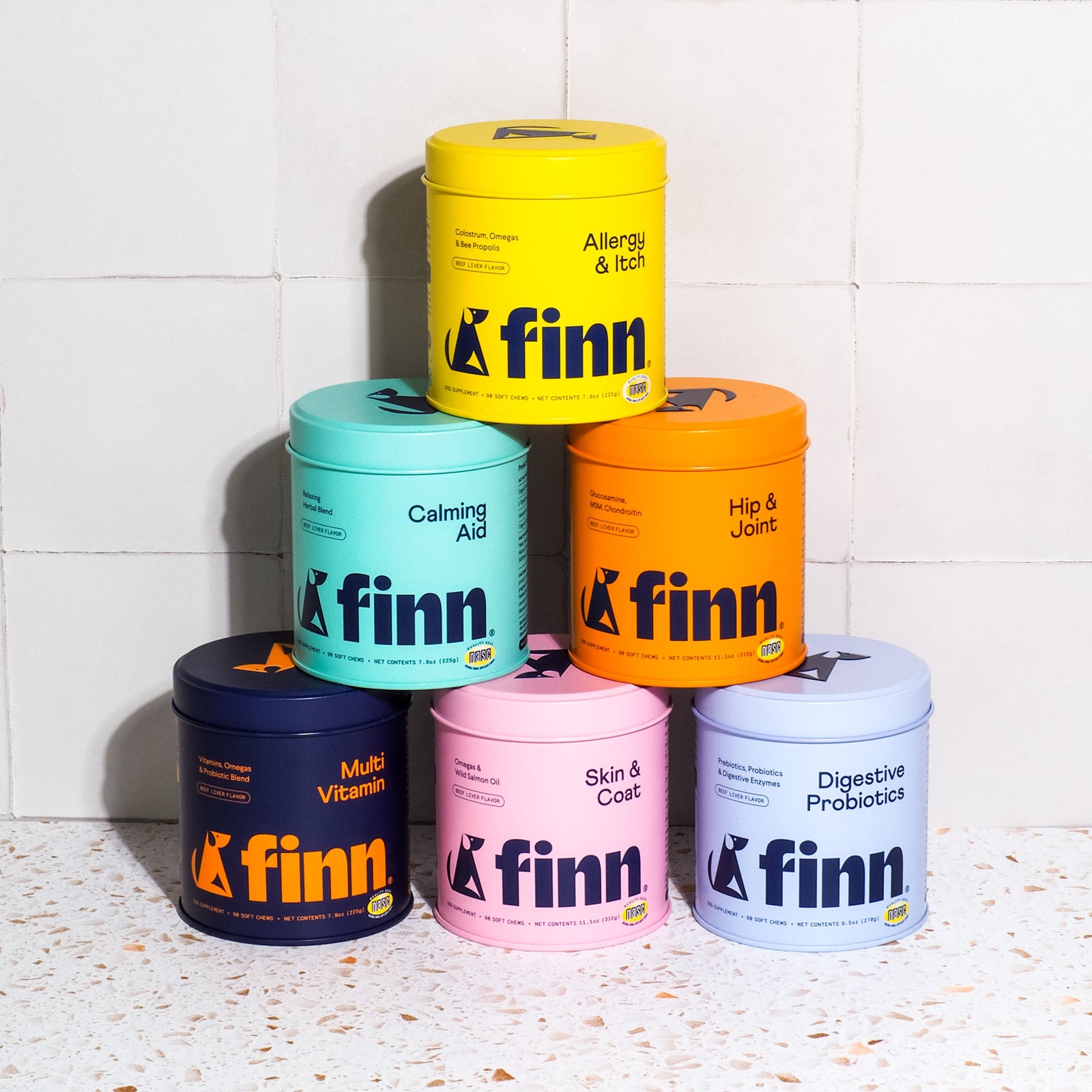 Six colorful Finn pet supplement containers stacked in pyramid formation, featuring yellow Allergy & Itch, teal Calming Aid, orange Hip & Joint, navy Multi Vitamin, pink Skin & Coat, and purple Digestive Probiotics.