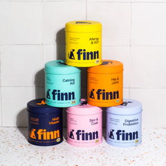 Six colorful cylindrical containers of Finn pet supplements stacked in a pyramid formation against white tile wall, featuring varieties like Allergy & Itch, Calming Aid, Hip & Joint, Multi Vitamin, Skin & Coat, and Digestive Probiotics in yellow, teal, orange, navy, pink, and light blue.