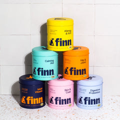 Six colorful cylindrical containers of Finn pet supplements stacked in a pyramid formation against white tile wall, featuring products for allergy, calming, joint, vitamin, skin, and digestive health.