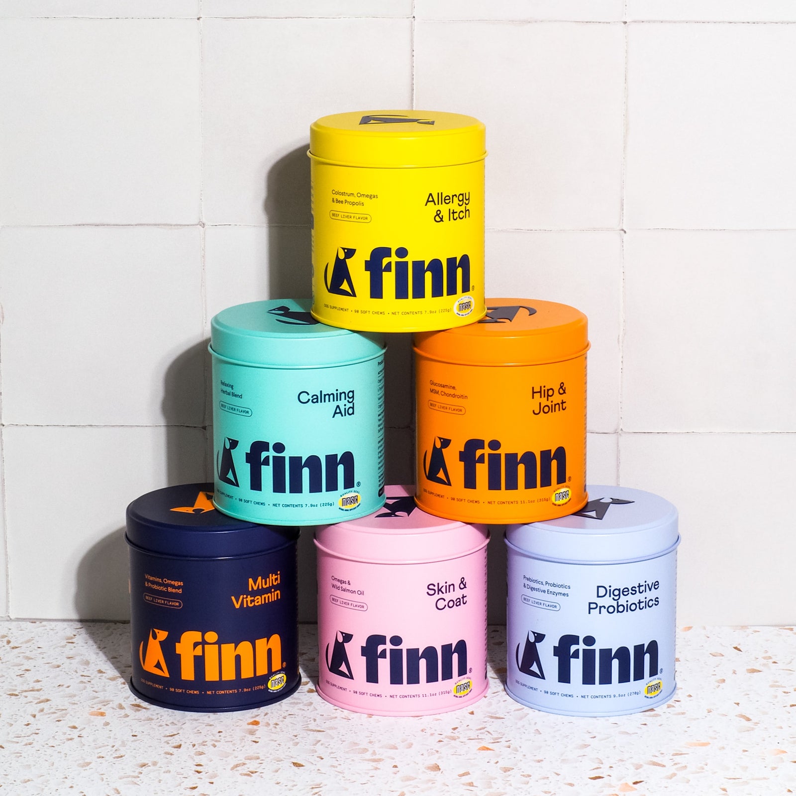 Six colorful Finn pet supplement containers stacked in pyramid formation against white tile wall - yellow, teal, orange, navy, pink, and purple canisters with different formulations like Allergy & Itch, Calming Aid, and Hip & Joint.