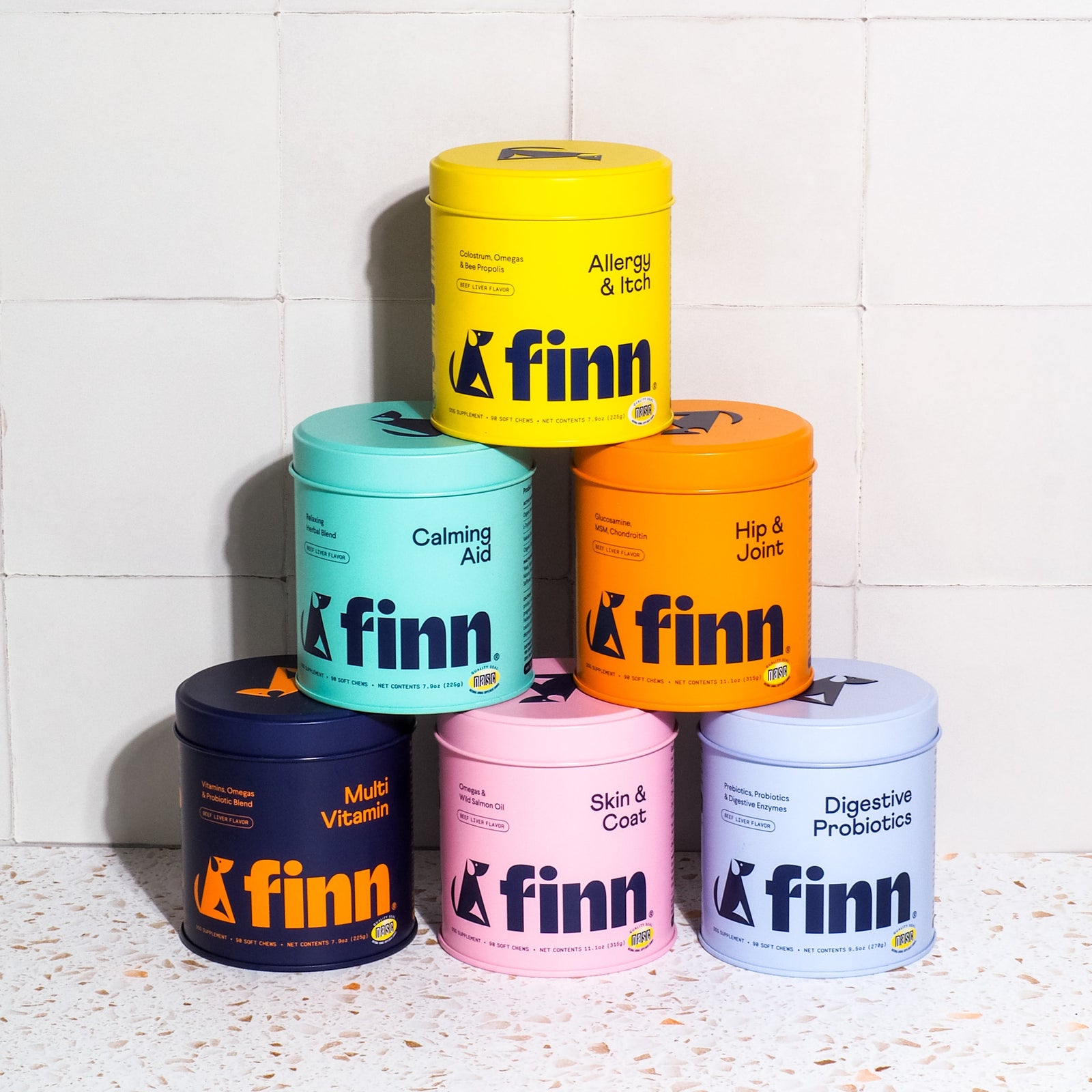 Six colorful cylindrical Finn pet supplement containers stacked in pyramid formation against white tile background, featuring yellow Allergy & Itch, teal Calming Aid, orange Hip & Joint, navy Multi Vitamin, pink Skin & Coat, and light blue Digestive Probiotics varieties.