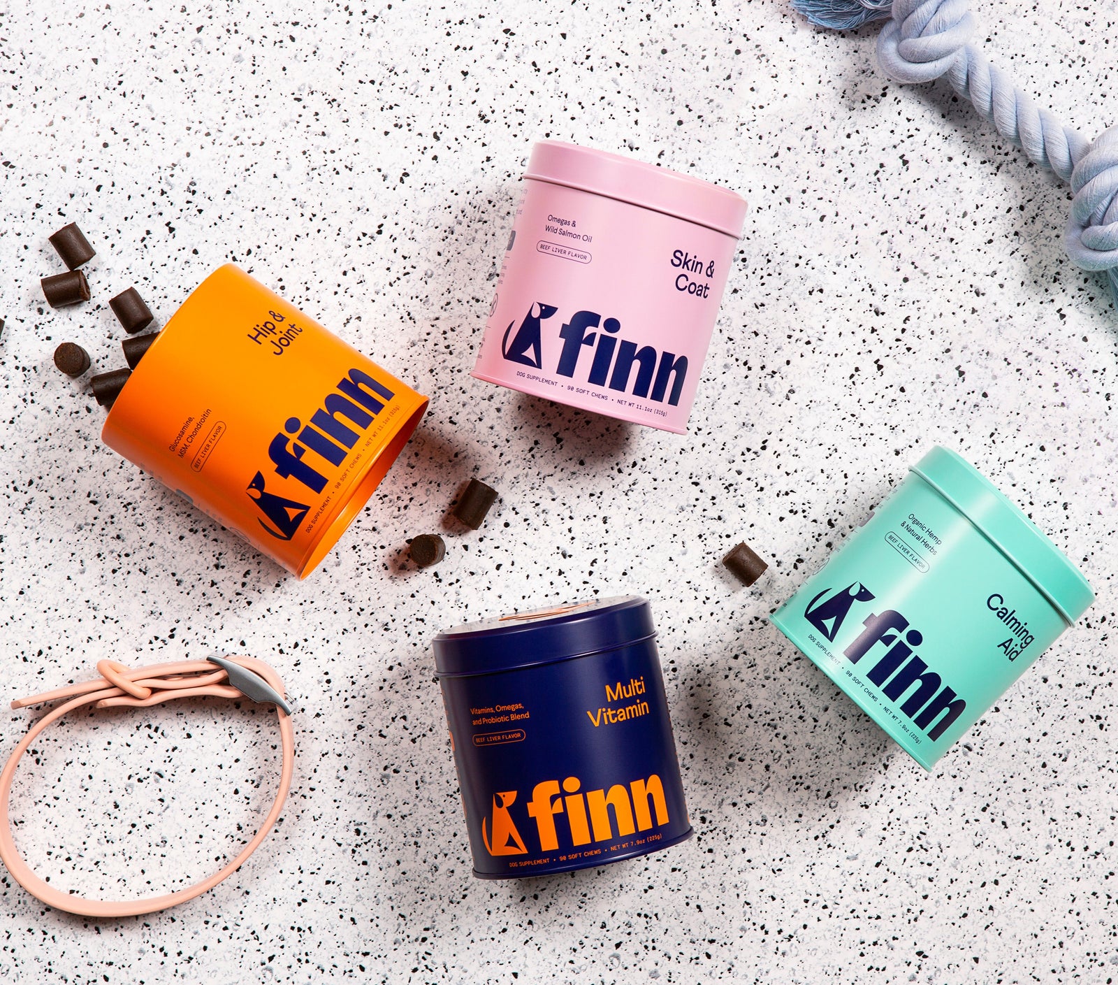Four colorful Finn supplement containers in orange, pink, navy, and mint green scattered on speckled white surface with chocolate pieces and rope details.