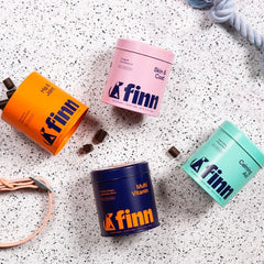 Four colorful Finn pet supplement containers arranged on speckled terrazzo surface with rope toy and treats scattered around